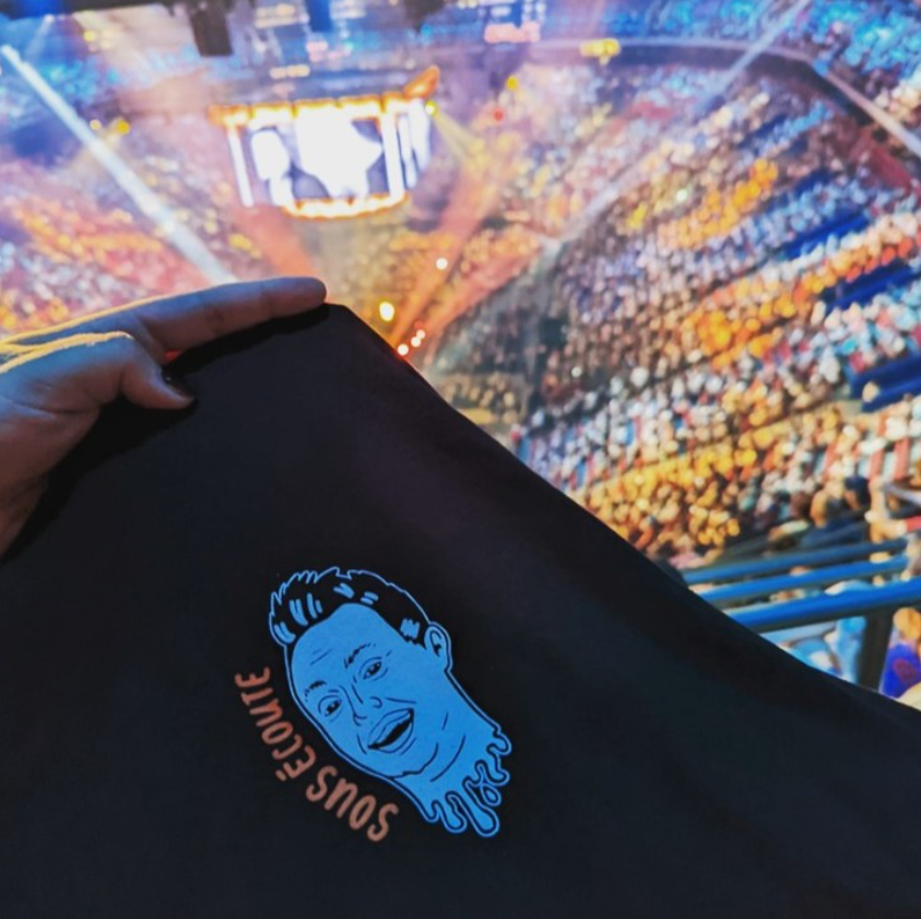 Mike Ward Sous Écoute black t-shirt with headless Mike logo. A filled arena can be seen behind.